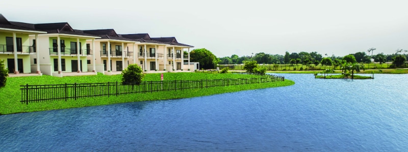 A tranquil view of the apartment complex is presented, surrounded by lush greenery and a serene body of water. The architecture features multiple balconies with a soft color palette, blending harmoniously with the landscape. A wooden fence borders the grassy area, enhancing the peaceful atmosphere.