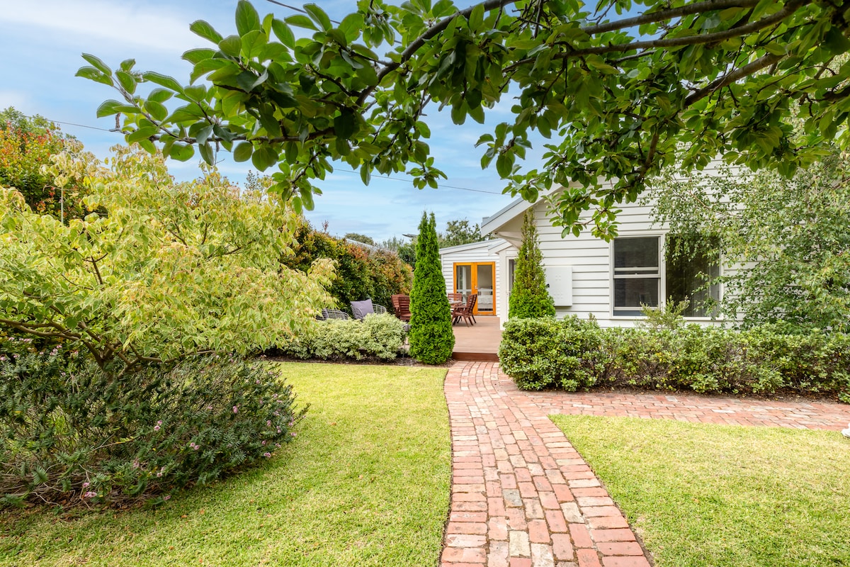 A charming pathway of brick leads to the front entrance of the house, bordered by manicured greenery. The yard features well-maintained shrubs and trees, with two outdoor seating areas visible beyond the entrance, inviting relaxation in a tranquil setting.
