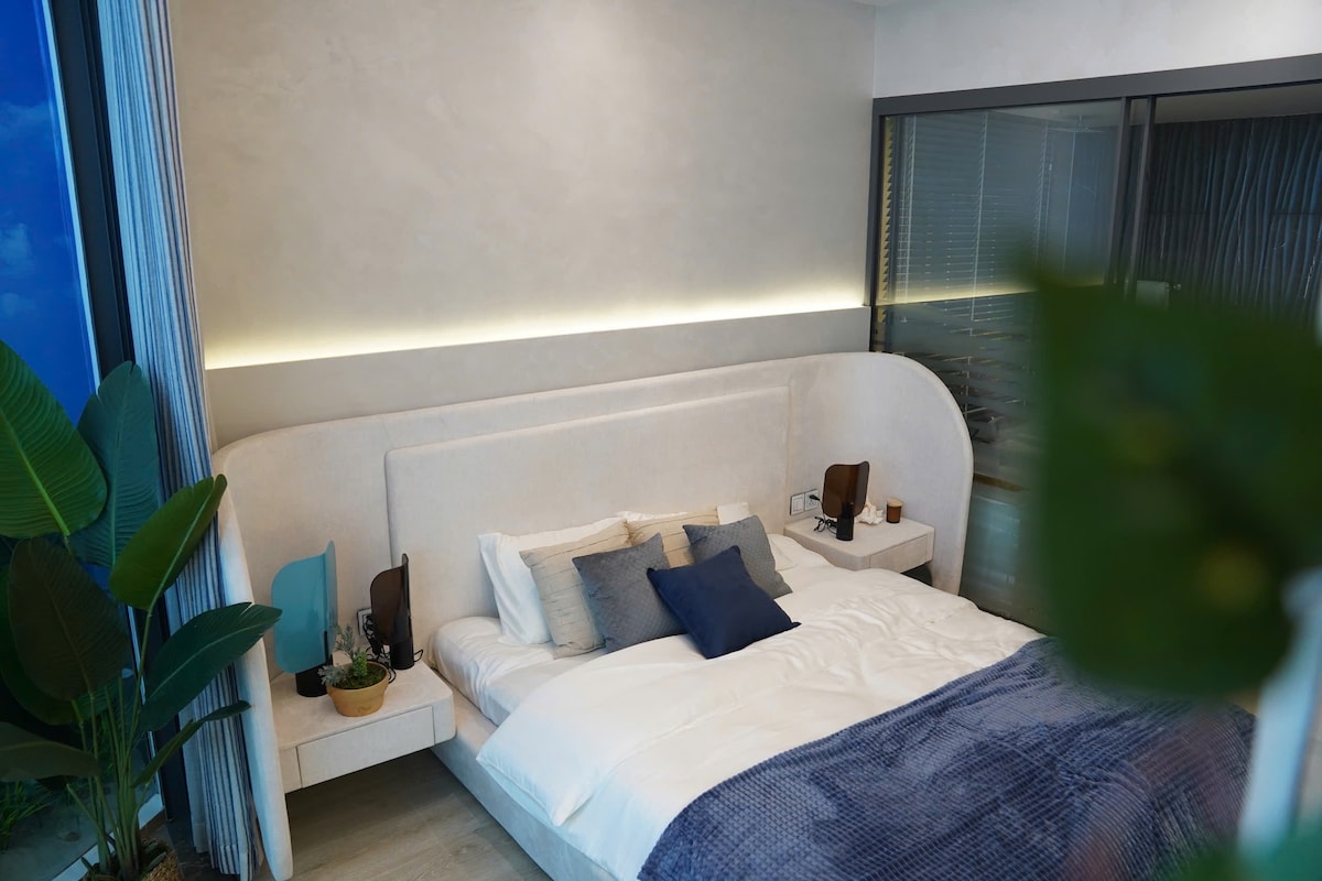 A modern sleeping area features a soft, curved headboard and a spacious bed layered with white and blue bedding. Nightstands are positioned on either side, complemented by decorative items. Green plants add a touch of nature, while ambient lighting creates a calm atmosphere.