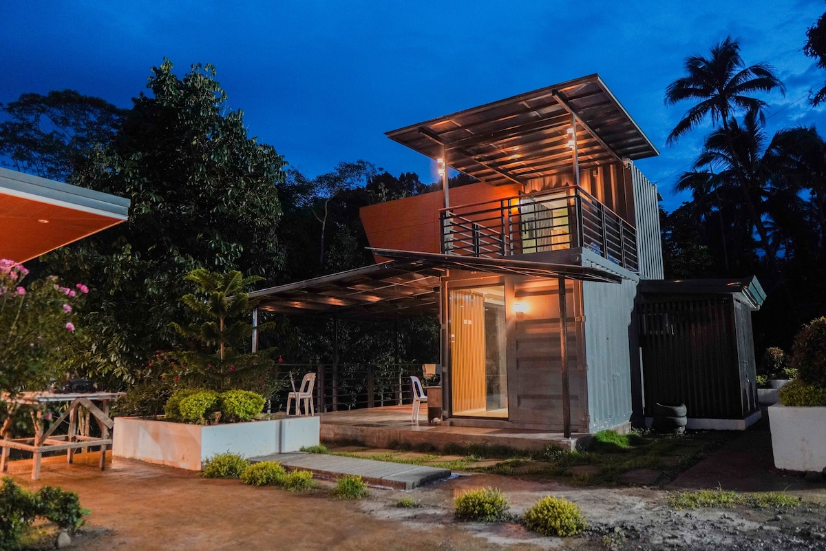 A modern container home is seen at twilight, showcasing a unique architectural design. Large windows and a balcony enhance the exterior, surrounded by landscaped greenery. Soft lighting highlights the entrance and outdoor space, creating an inviting atmosphere for guests arriving.