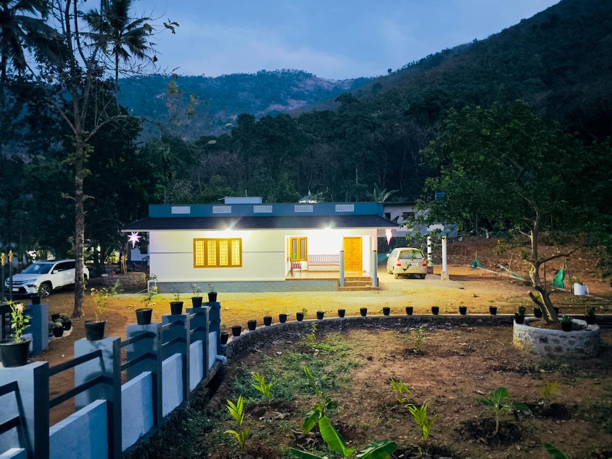 The spacious countryside retreat is framed by lush greenery and mountains. Soft light emanates from the windows, illuminating the exterior. A neatly arranged garden, featuring potted plants and a circular path, leads to the entrance, complementing the tranquil ambiance of the setting.