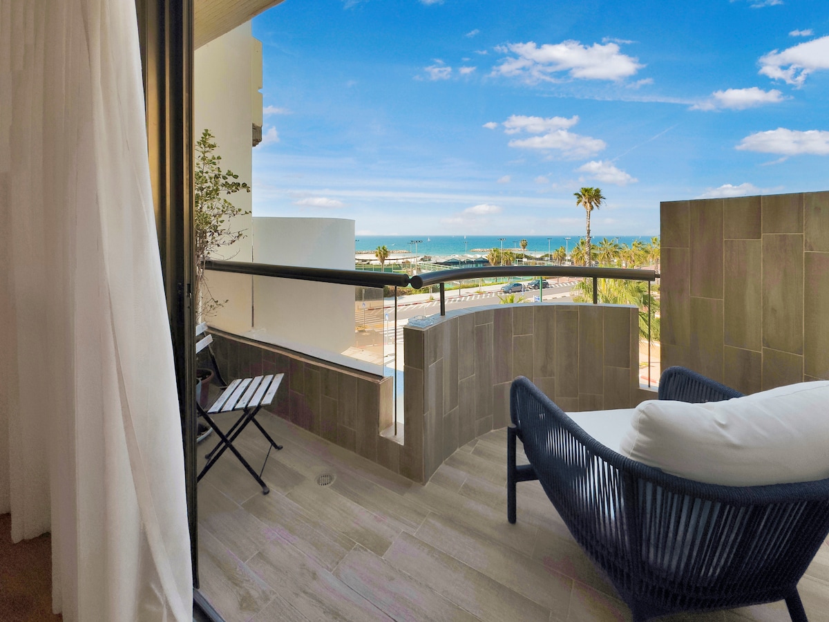 A private balcony offers views of the coastline, with the sea visible beyond palm trees. Comfortable outdoor furniture is present, including a lounge chair and a small table. The balcony is bordered by glass railing, allowing for unobstructed scenic enjoyment.