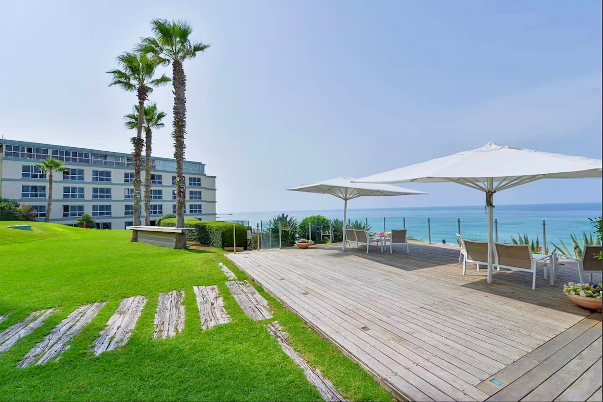 A spacious beachfront deck showcases wooden flooring and is surrounded by lush green grass. Two umbrellas provide shade over lounge chairs, with palm trees gently swaying nearby. The soothing view of the Mediterranean Sea can be seen in the background, enhancing the outdoor relaxation space.