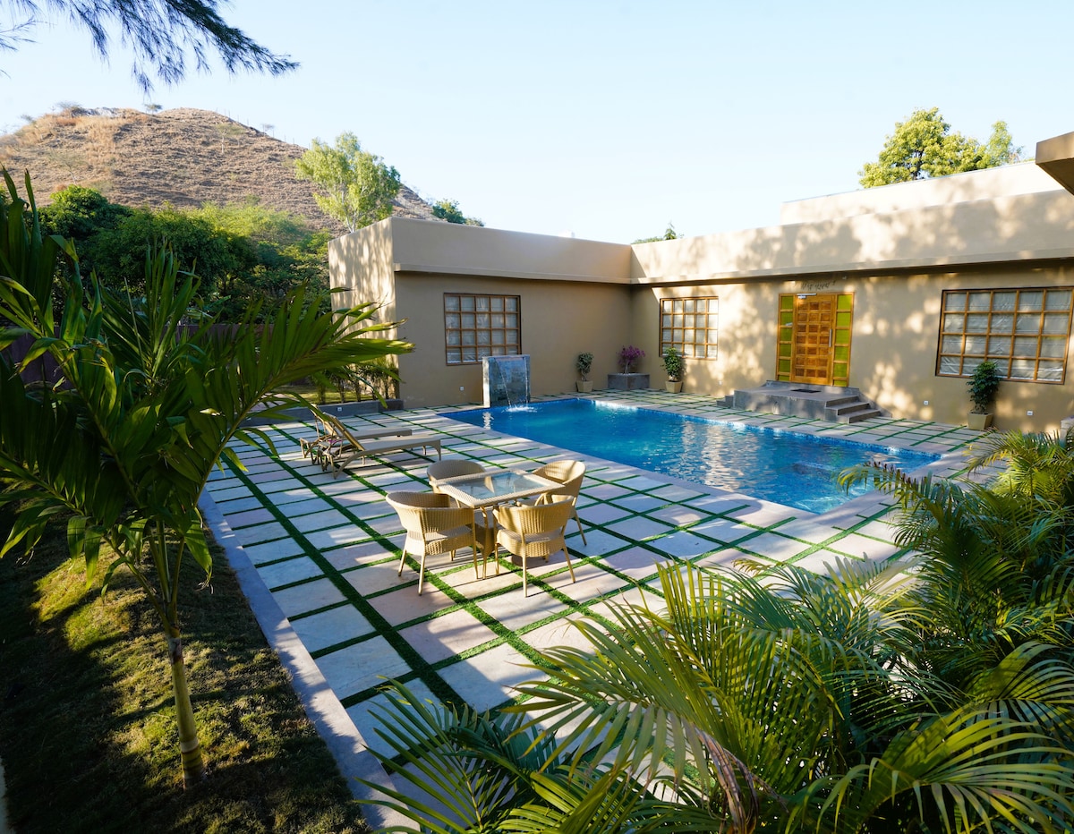 A serene outdoor area features a swimming pool surrounded by well-maintained greenery and stone pathways. Comfortable lounge chairs are positioned near the pool, inviting relaxation. The backdrop includes a gentle hillside and the structure's large windows, enhancing the connection with nature.