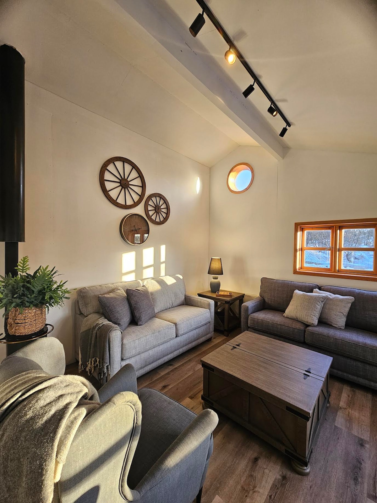 A cozy living room features two gray sofas and a stylish coffee table, complemented by a small lamp and decorative items. Natural light filters through the circular window and nearby framed window, creating a warm ambiance. Rustic accents, including wall décor, contribute to the inviting atmosphere.