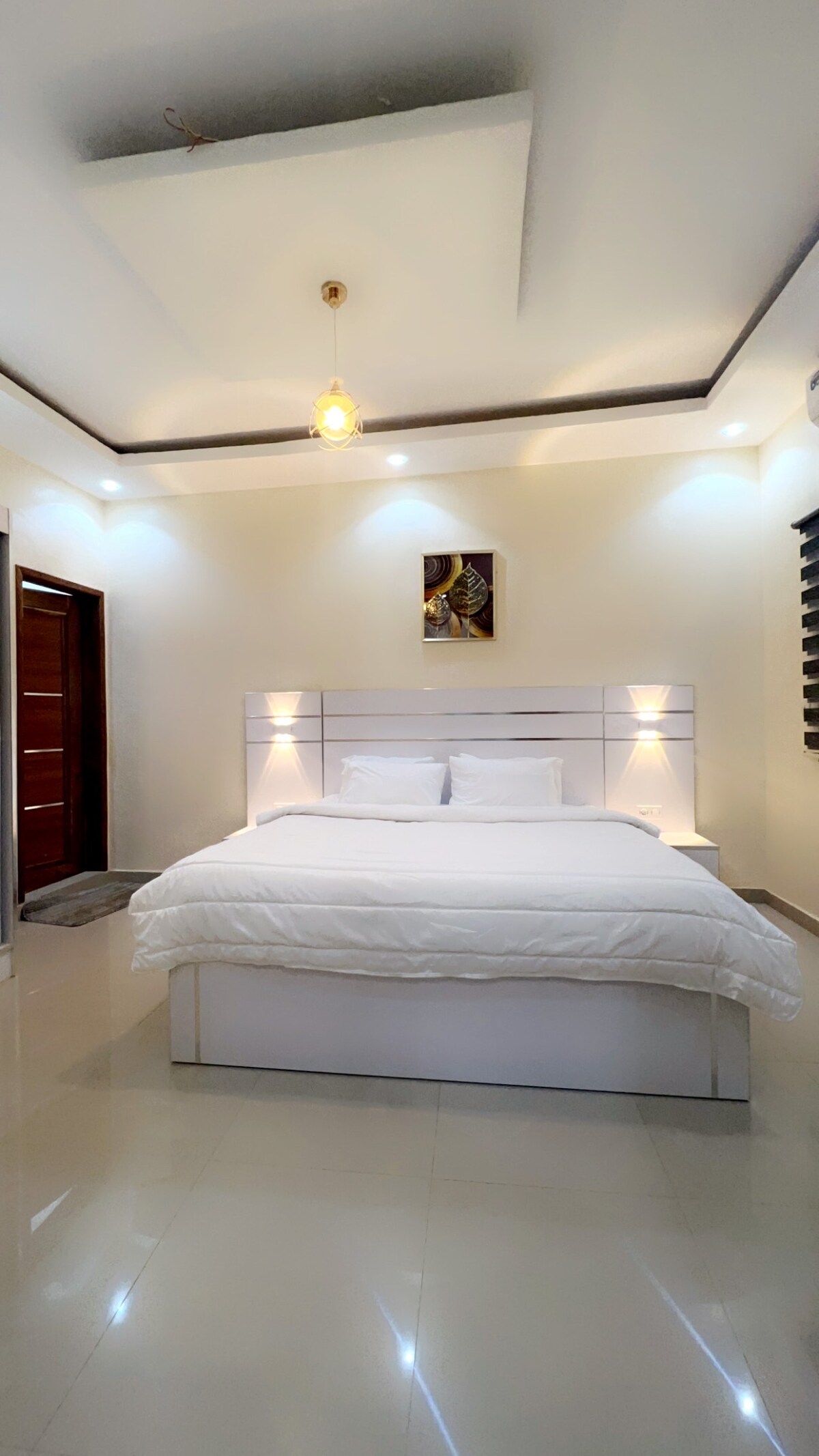 A well-lit bedroom features a spacious bed with white bedding and two pillows. Soft lighting from bedside fixtures enhances the room's modern design. A decorative wall piece adds an artistic touch, while sleek wooden doors are visible on the left side.