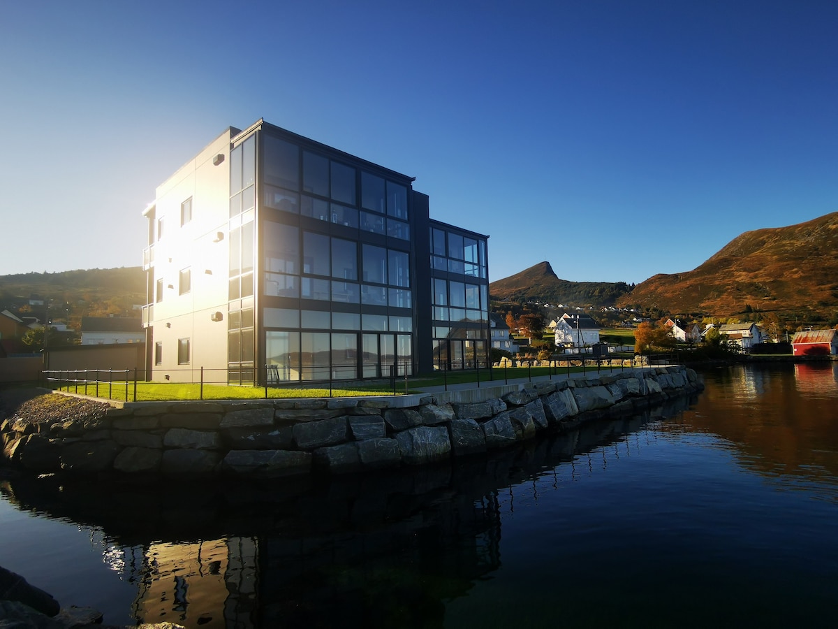 A modern building with multiple glass panels reflects sunlight, surrounded by a serene water surface. The structure is set against rolling hills, enhancing the natural landscape. An expansive grassy area is visible in front, offering a sense of openness and connection to the surroundings.