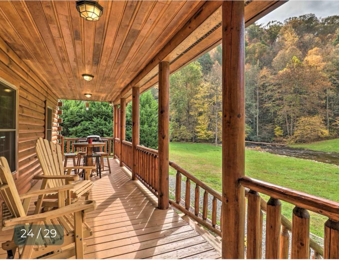 A spacious wooden porch features several Adirondack chairs, providing a comfortable seating area. The railing adds a rustic touch. Lush green landscaping surrounds the cabin, and trees with autumn foliage are visible beyond the porch, suggesting a serene natural environment.