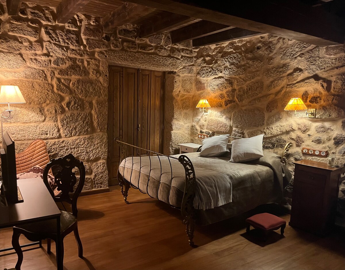 A cozy bedroom features stone walls and wooden beams, enhancing its rustic charm. A spacious bed with soft linens is placed against one wall, while warm light is provided by two bedside lamps. A vintage desk and an ornate chair add to the inviting decor.
