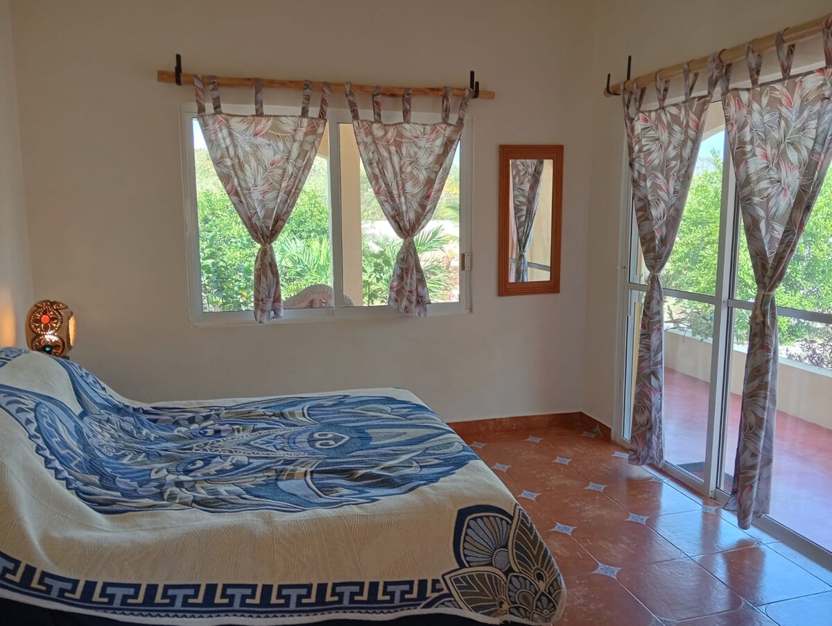 A bright bedroom features a spacious bed adorned with a patterned blanket. Natural light enters through large windows lined with patterned curtains. A decorative mirror is mounted on the wall, and views of greenery are visible outside.