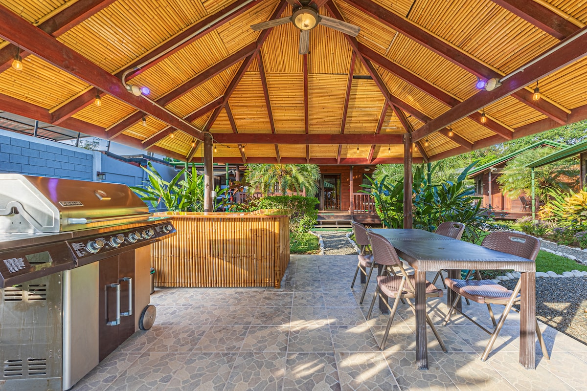 An outdoor patio area features a wooden ceiling with exposed beams above a spacious dining table and four chairs. A gas grill is positioned nearby, and lush greenery surrounds the space, creating a tranquil atmosphere for outdoor dining and relaxation.