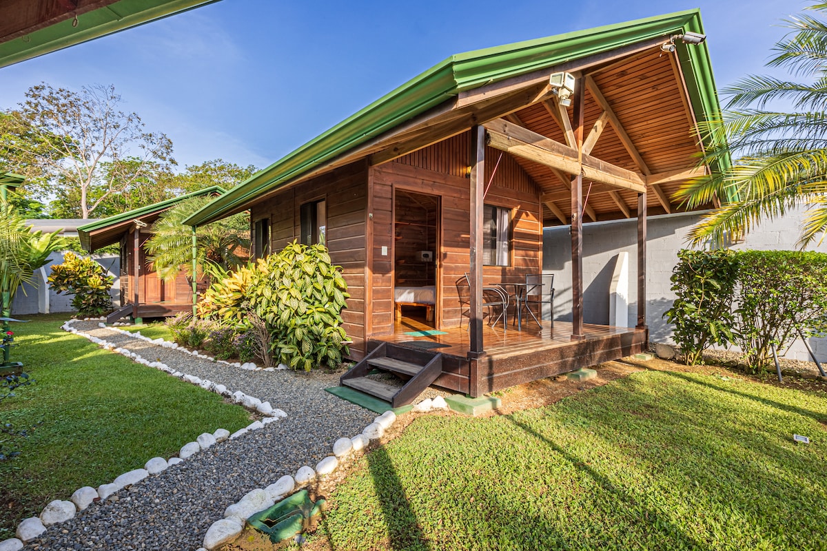 A wooden bungalow is set against a clear blue sky, showcasing a welcoming front porch with chairs. Lush greenery surrounds the structure, complemented by neatly arranged stone pathways and vibrant tropical plants. The sloped roof features a green overhang, adding character to the retreat.