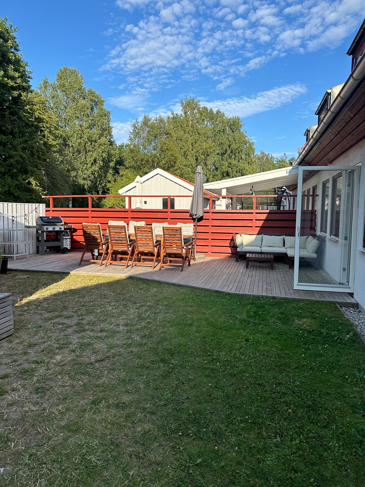 The outdoor patio features a dining area with several wooden chairs arranged around a table. A grill is positioned nearby, and an umbrella provides shade. Adjacent to the house, large windows allow natural light into the living space, surrounded by trees for a tranquil environment.