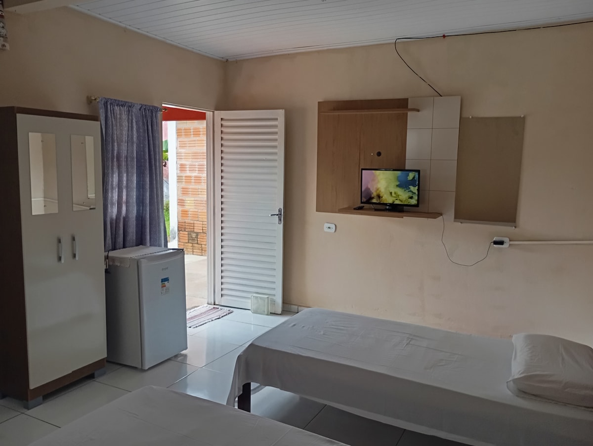 The room features a simple layout with two single beds and a small wall-mounted TV. A mini-fridge is positioned next to a two-door wardrobe. Natural light enters through the open door, which leads to the outdoor area.