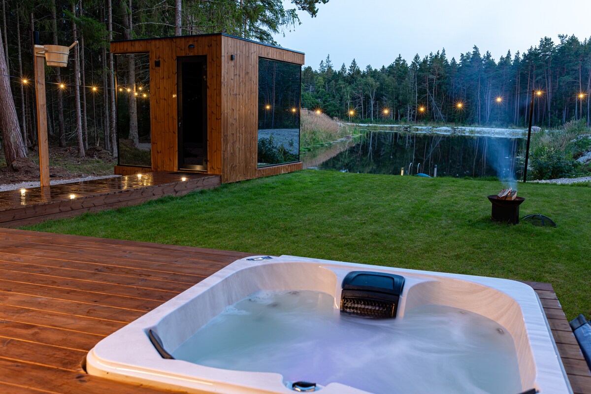 A hot tub is positioned on a wooden deck, overlooking a serene body of water. In the background, a wooden sauna stands beside well-maintained grass, with warm outdoor lights strung overhead, adding a relaxed ambiance to the natural setting.