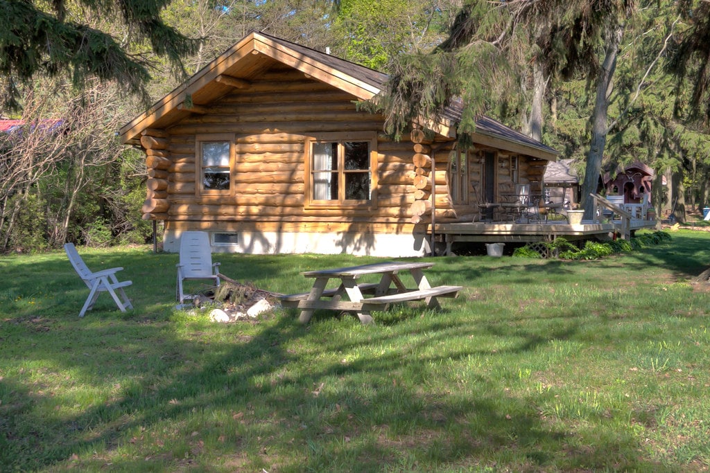 The log cabin is set on a grassy area surrounded by trees, featuring a picnic table and a couple of outdoor chairs arranged for relaxation. The cabin's rustic exterior is complemented by windows that provide natural light.
