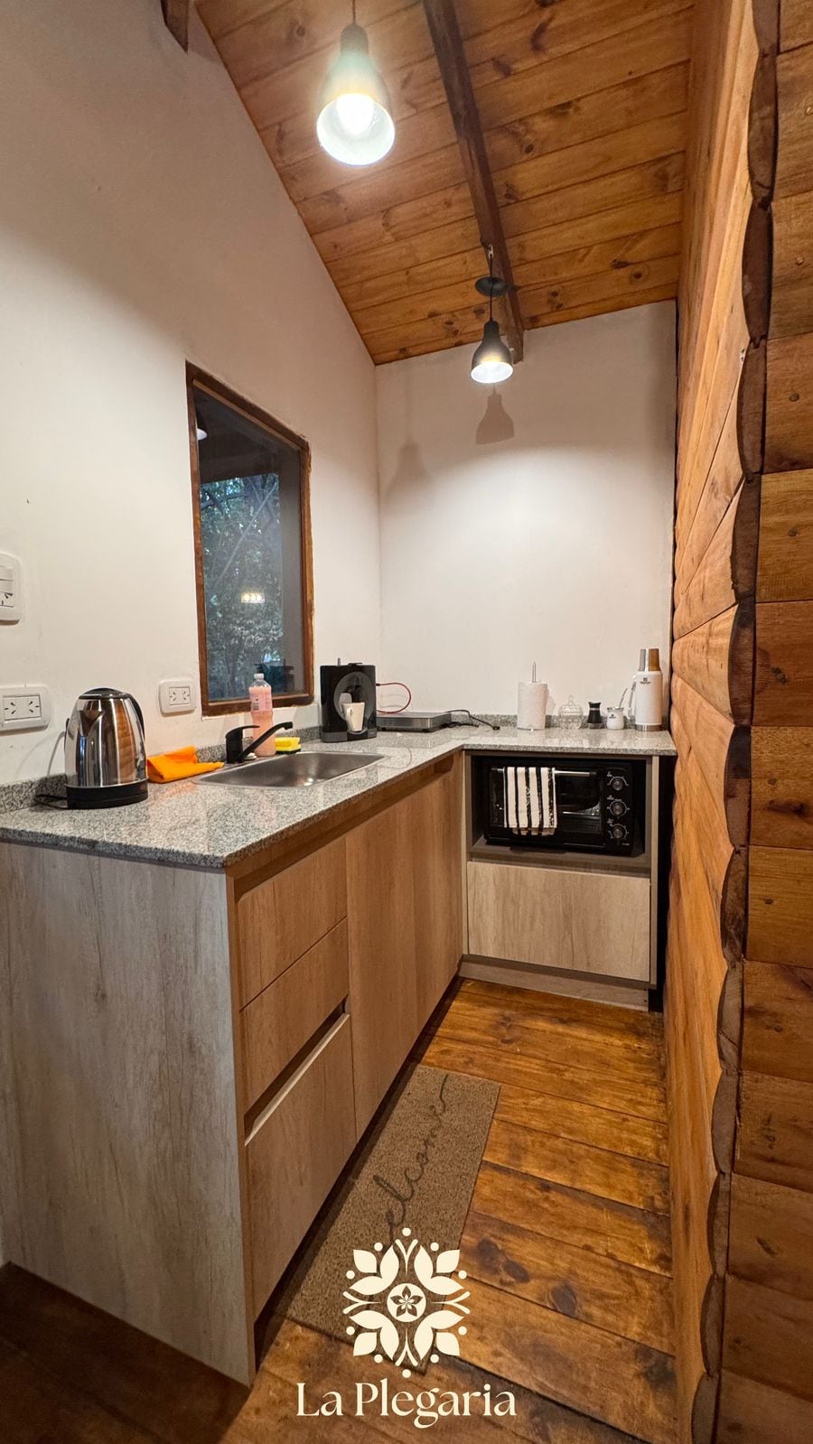 A compact kitchen area features wooden accents and modern appliances. A granite countertop offers ample space for meal preparation, while a kettle and coffee maker are visible. Wall-mounted lights provide illumination, and a window allows natural light to enhance the warm wooden tones of the space.