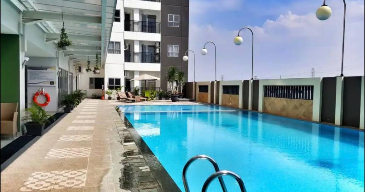 An inviting pool area is featured with clear blue water, surrounded by landscaped spaces and lounge chairs. Modern lighting fixtures line the poolside, illuminating the relaxing environment. The building's sleek exterior is visible in the background against a clear sky.