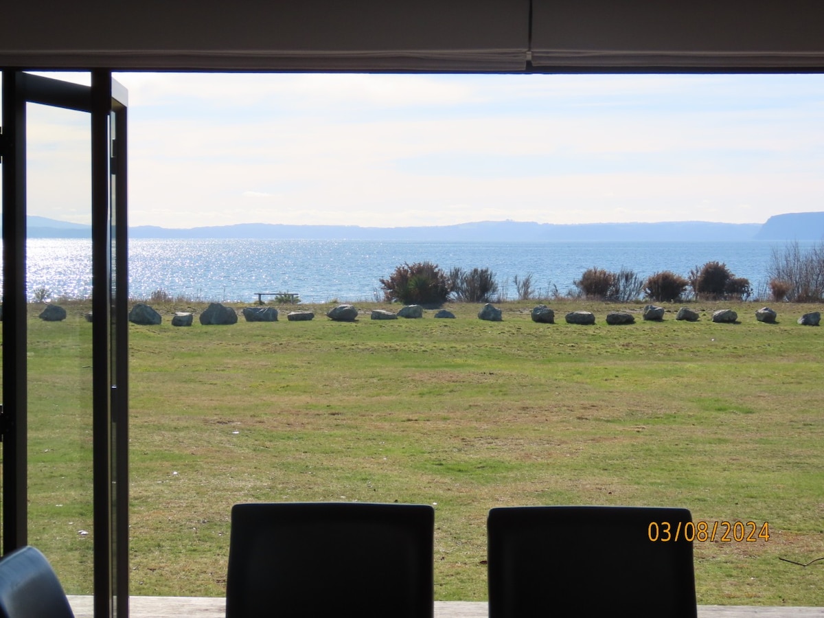 A scenic view of a tranquil lake is captured from the open doorway, where a grassy area leads to the water's edge. Stones are arranged along the shoreline, with distant picnic areas visible under a clear sky. Natural light brightens the scene.