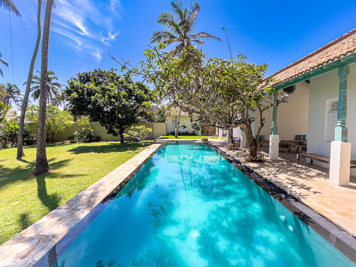 A tranquil private pool is surrounded by a spacious garden, featuring lush greenery and tall palm trees. Sunlight reflects off the clear water, creating a calm ambiance. The pool is complemented by a stone border, enhancing the outdoor space.