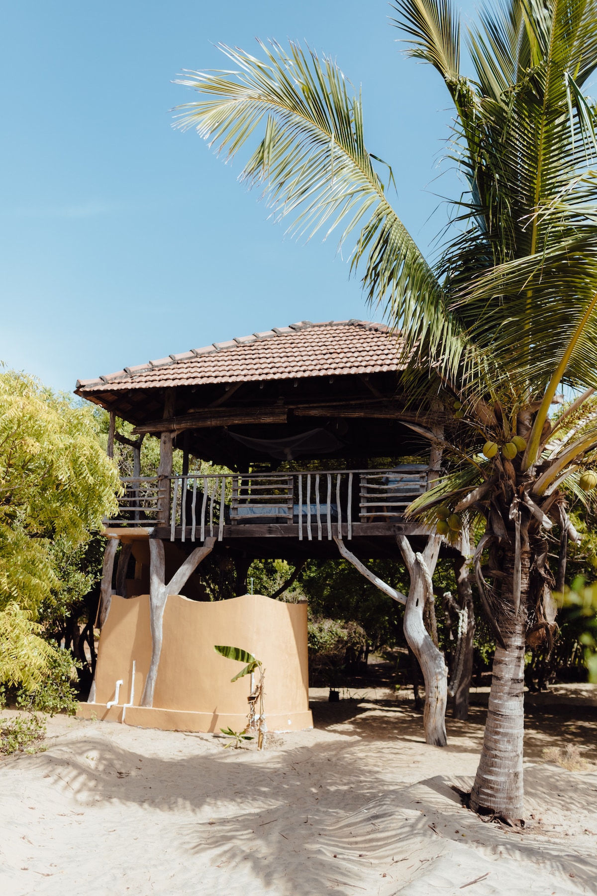 A unique wooden cabana is elevated on stilts surrounded by lush greenery and sandy ground. The structure features a thatched roof, open balcony railings, and natural wood accents, offering a tropical retreat under a clear blue sky.