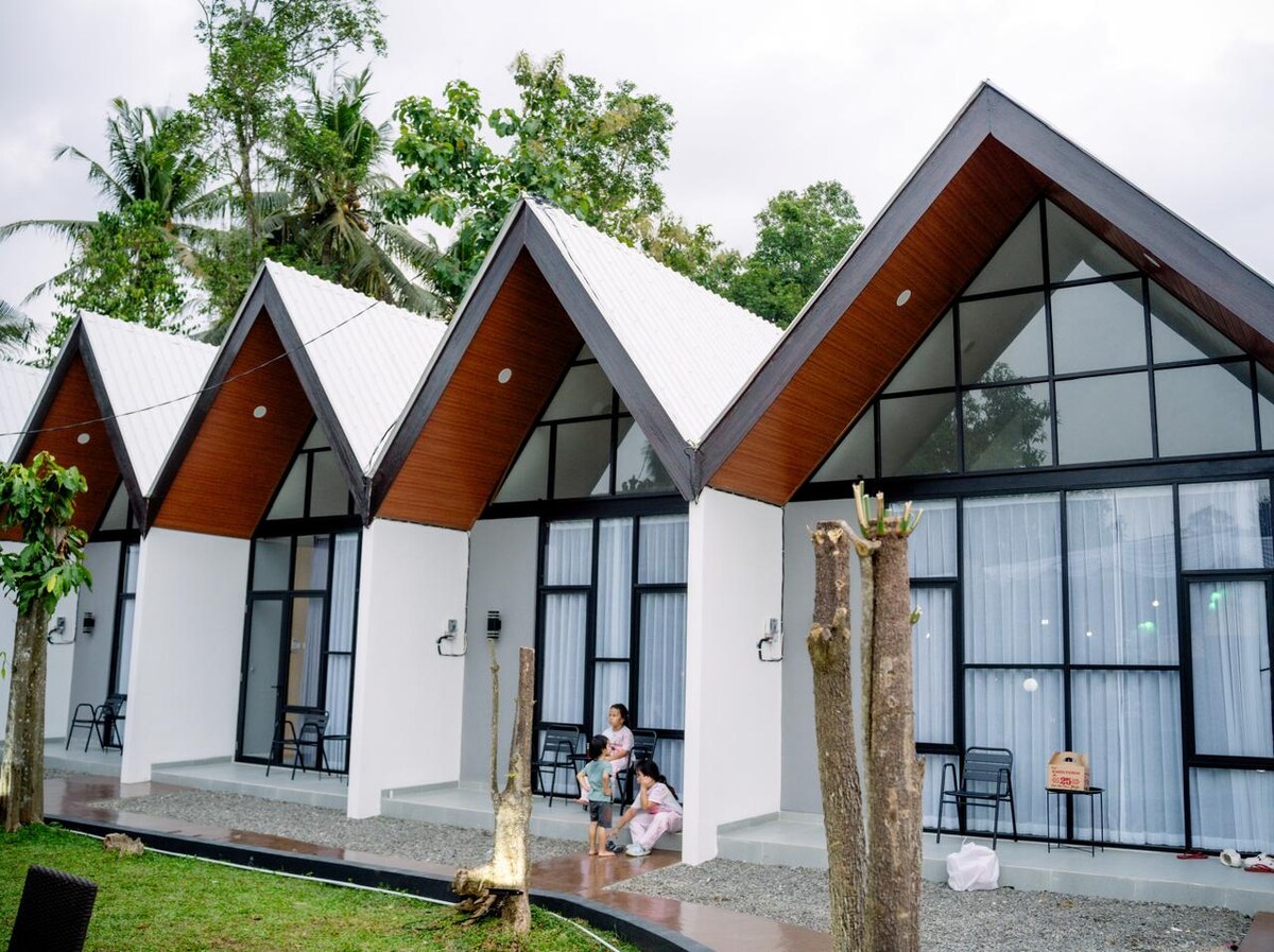 A row of modern bungalows is set amidst lush greenery, featuring sloped roofs and large windows. Each bungalow has an outdoor seating area with chairs, enhancing the tranquil ambiance. The pathways are surrounded by well-maintained landscaping, contributing to a peaceful retreat.