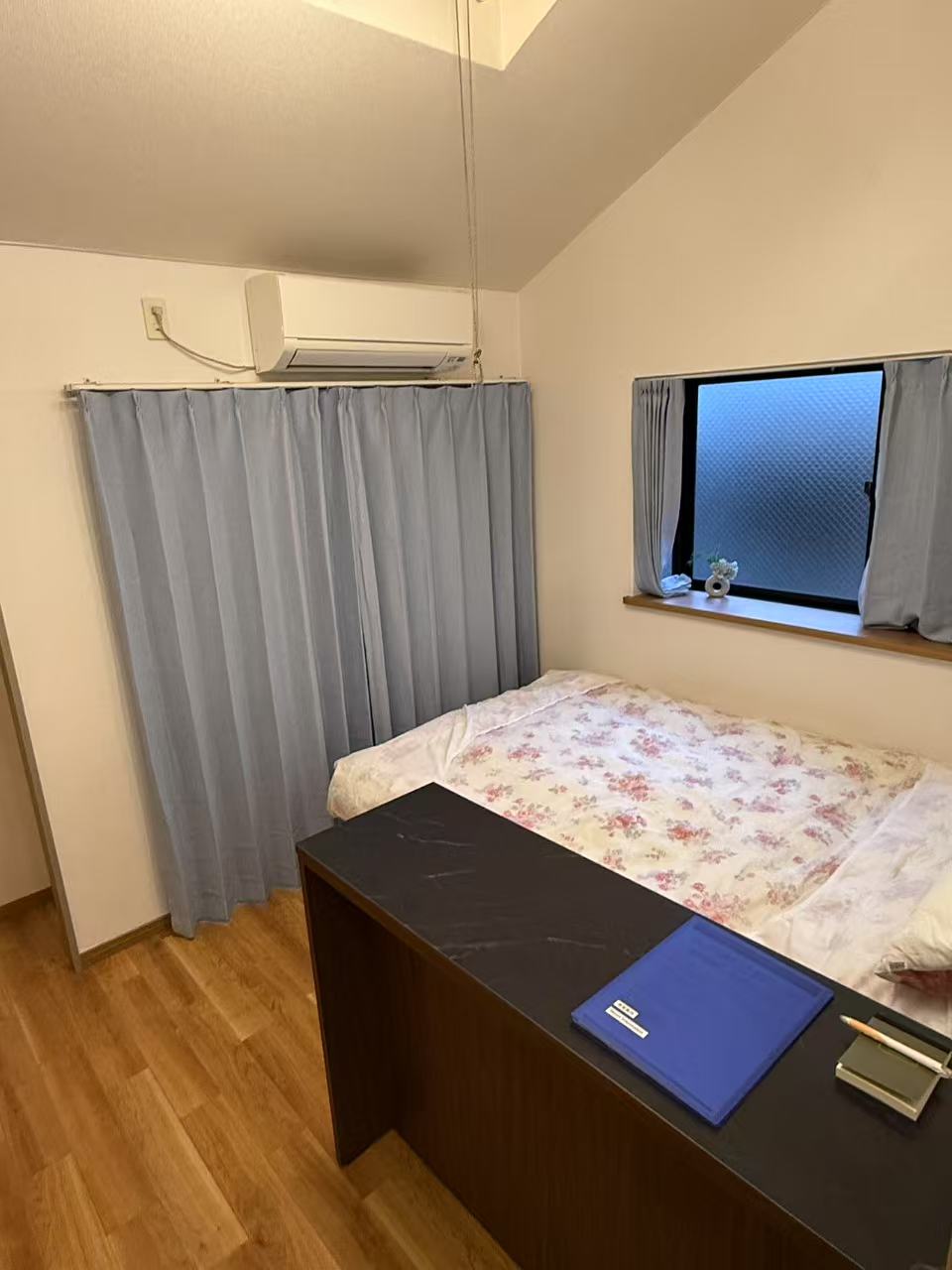 A bedroom features a spacious bed dressed with floral bedding. A window is partially covered by light blue curtains, allowing natural light to filter in. An air conditioning unit is mounted above the bed, and a desk with stationery is situated nearby.