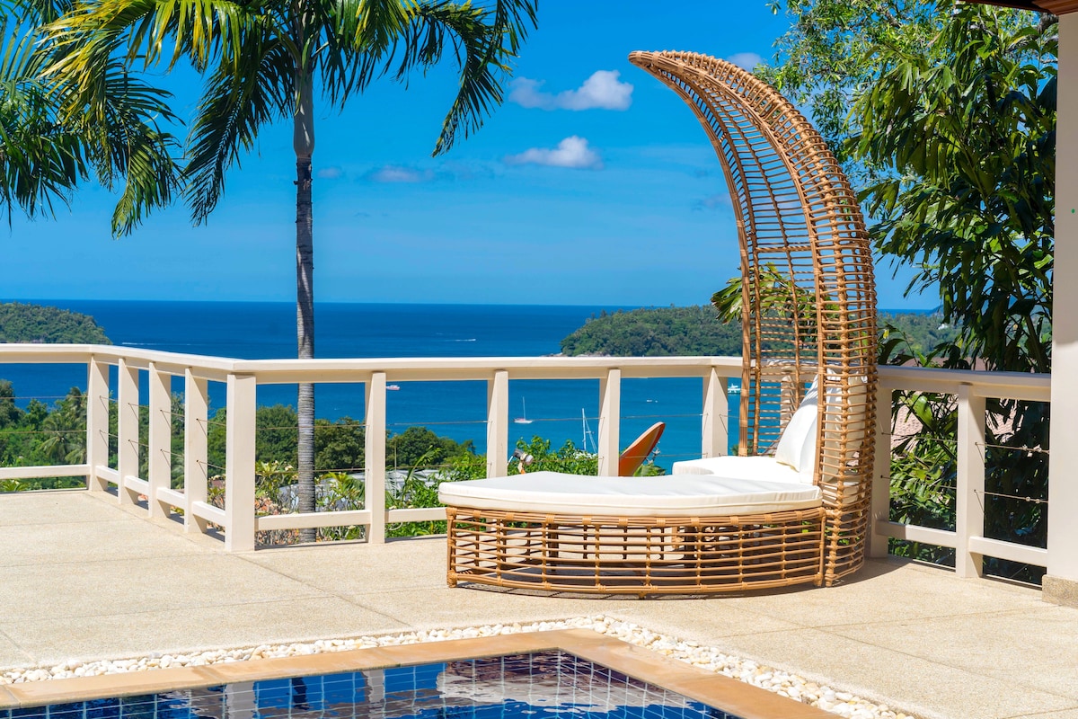 A stylish lounge chair made of natural materials is positioned by the pool, overlooking the expansive view of the Andaman Sea and lush greenery. The clear blue sky complements the serene atmosphere, inviting relaxation in this tranquil outdoor space.
