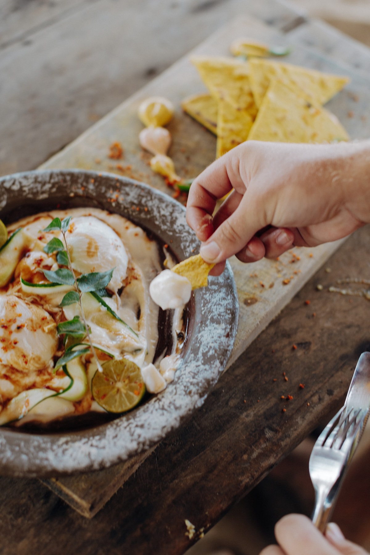 A hand reaches for a tortilla chip, lightly dipped in creamy sauce, from a traditional dish presented on a rustic wooden table. Surrounding elements include sliced cucumbers, lime wedges, and a sprinkle of spices, creating an inviting and vibrant meal setting.