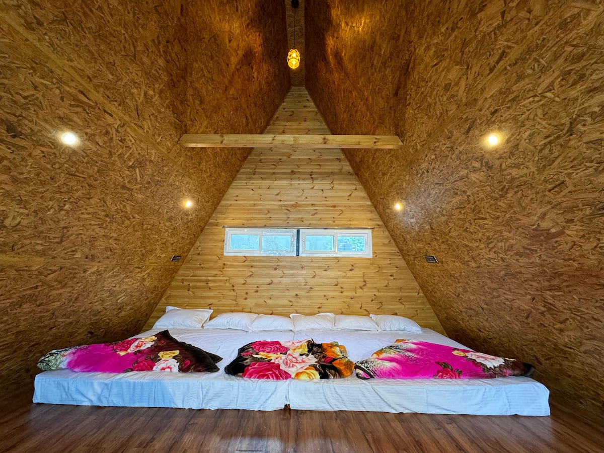 An inviting sleeping area features a spacious arrangement of bedding on a wood floor, complemented by colorful blankets with floral patterns. The triangular wood-paneled walls enhance the cozy atmosphere, while natural light filters through small windows near the top, illuminating the room.