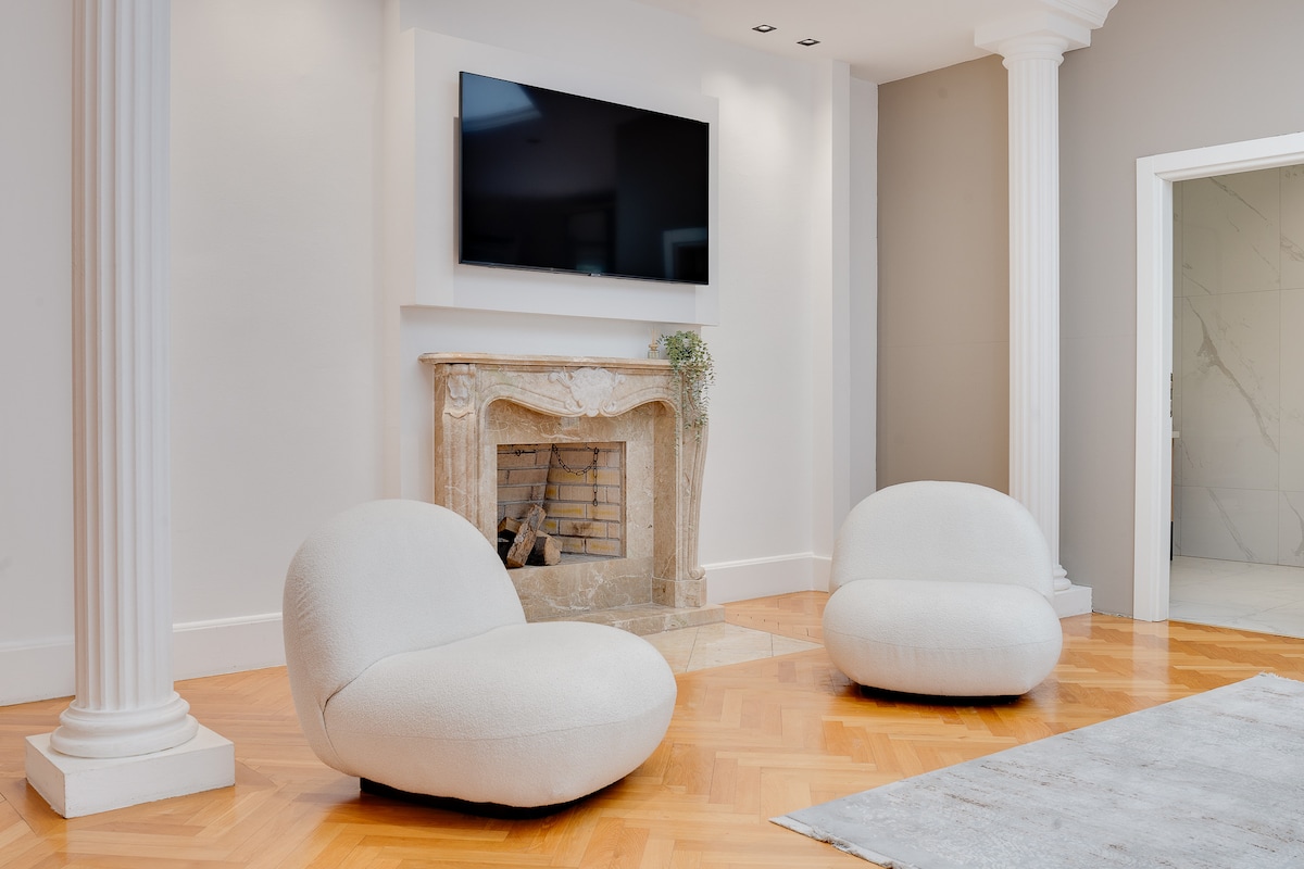 A well-lit lounge area is shown, featuring two soft, rounded chairs positioned on a polished wooden floor. A modern fireplace with a stone surround is visible, complemented by a wall-mounted television above. Elegant pillars frame the space, enhancing the room's design.