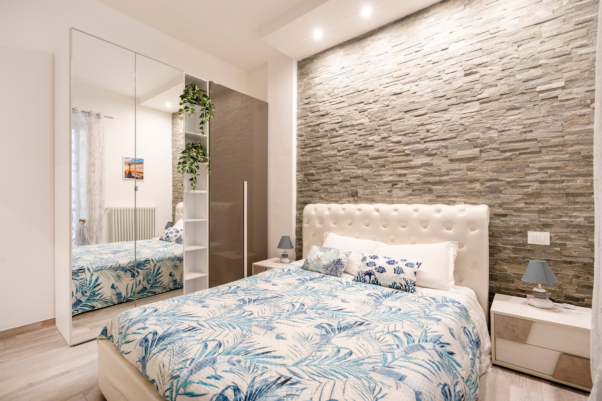A serene bedroom features a spacious bed with a light-colored tufted headboard and a patterned blue and white bedspread. A mirrored wardrobe reflects natural light, while a soft lamp illuminates two nightstands on either side. A plant adds a touch of nature to the decor.