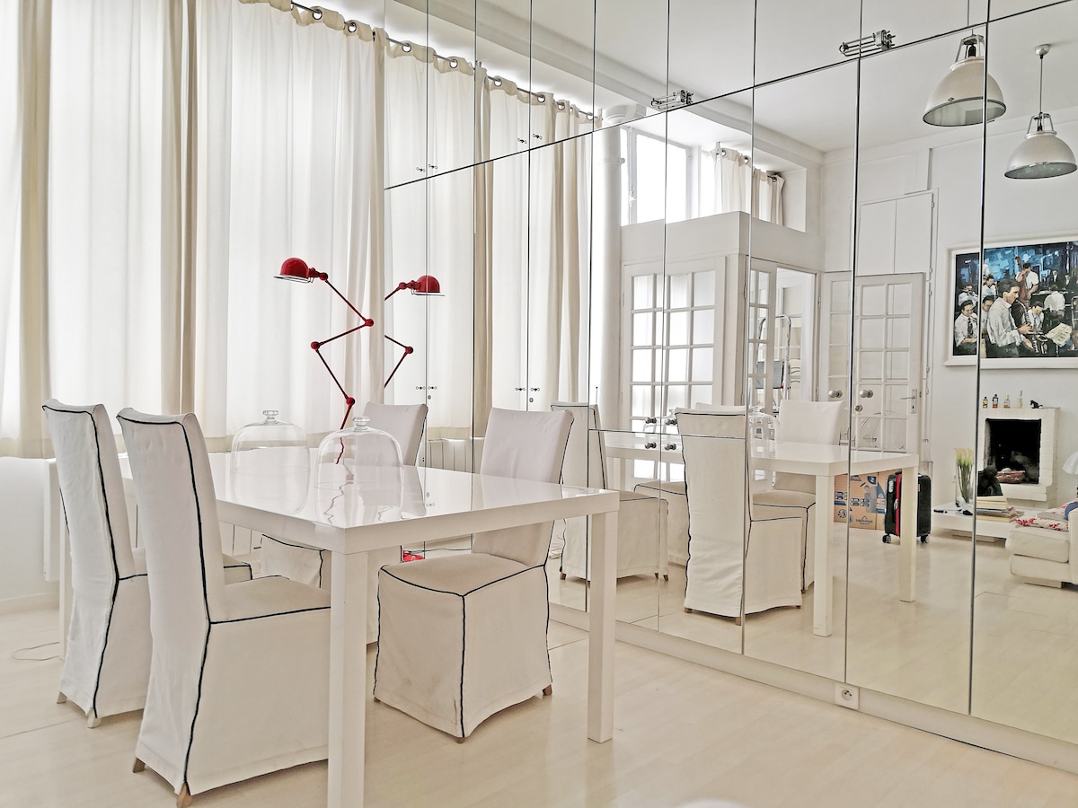 A bright dining area is presented, featuring a white table surrounded by several upholstered chairs. Large mirrors line one wall, reflecting natural light from tall windows, which are draped with sheer curtains. Minimalist pendant lights hang above, adding to the contemporary decor.