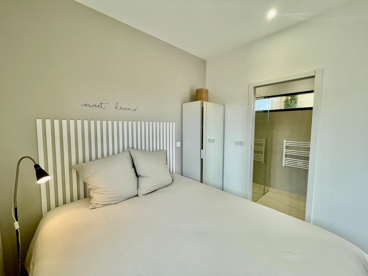 A serene bedroom features a spacious bed with neutral linens and soft pillows. A minimalistic bedside lamp stands beside the bed, while a wardrobe subtly complements the room's decor. An ensuite bathroom can be seen through an open doorway, enhancing the room’s functionality.