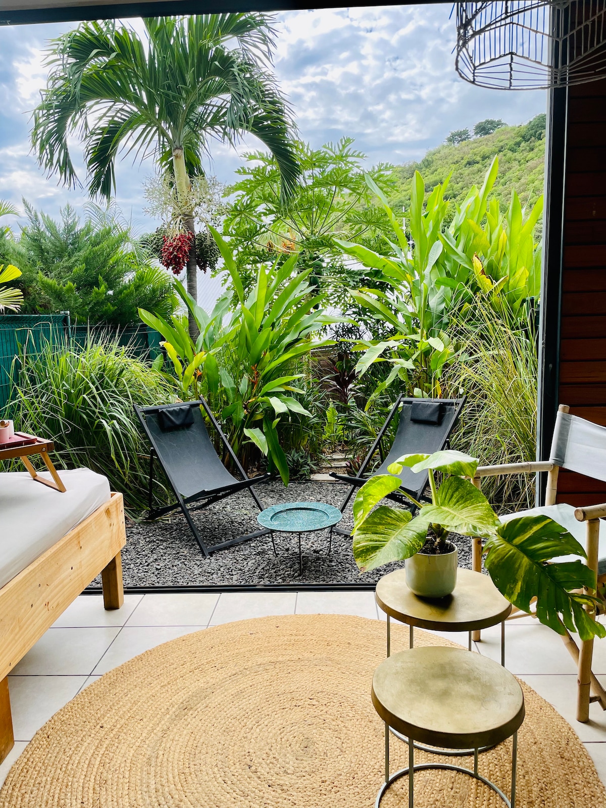 An outdoor seating area is framed by lush tropical plants and vibrant greenery. Two black lounge chairs are positioned around a small circular table, while a round natural fiber rug sits beneath. A small potted plant adds a touch of color to the setting.
