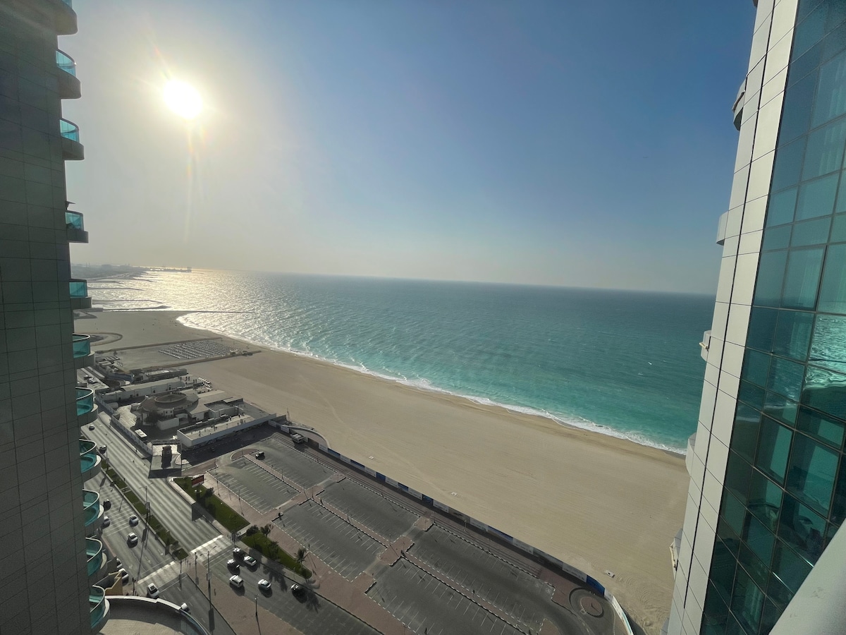 A panoramic view of the ocean is captured from the large balcony, showcasing sparkling turquoise water meeting a sandy beach. The sun casts a warm glow over the scene, and tall buildings frame the edges, creating a sense of elevation.