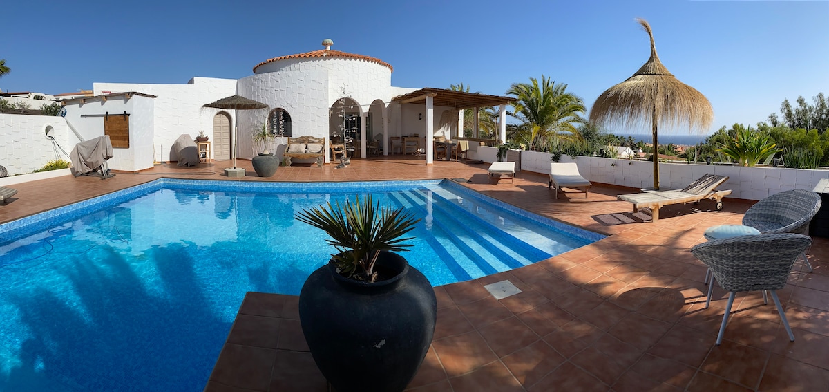 A spacious outdoor area features a private pool surrounded by sun loungers and palm trees. A thatched-roof canopy provides shade, while a dining area is visible in the background. The clean blue water of the pool reflects the clear sky, offering a serene ambiance.