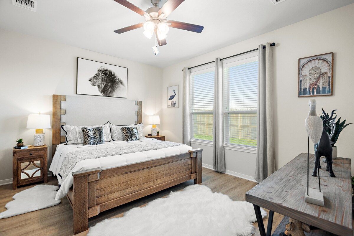 A serene bedroom features a spacious bed with a soft white cover and decorative pillows. Two bedside tables hold lamps, and a wooden dresser is positioned nearby. Natural light enters through two windows, framed by light gray curtains, illuminating the pale wooden flooring.