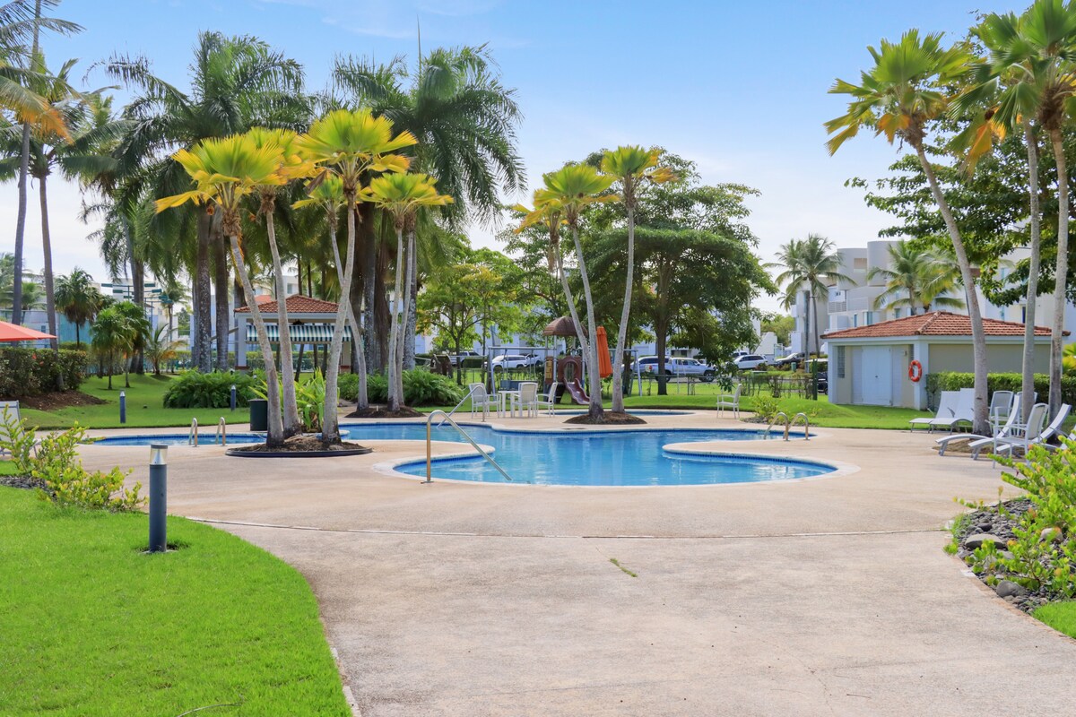 A serene pool area is surrounded by tropical palm trees and manicured lawns. The crystal-clear water invites relaxation, while lounge chairs are positioned for sunbathing. Pathways lead to nearby facilities, emphasizing a tranquil atmosphere in the vibrant outdoor setting.