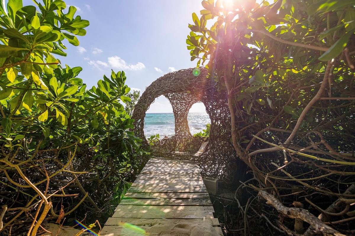 A wooden path winds through lush greenery, leading towards a natural archway made of intertwined branches. The sun filters through the foliage, casting dappled light on the sandy surface. Beyond, the tranquil sea is visible, inviting exploration and relaxation.