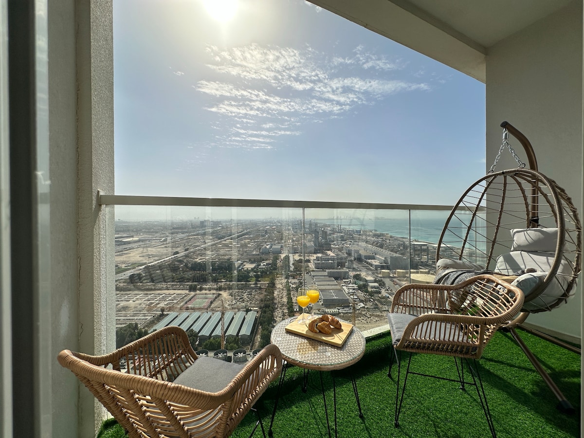 A balcony space features a hanging chair and two wicker chairs surrounding a small table set with refreshments. The balcony offers expansive views of the city and the sea, framed by clear skies and bright sunshine.