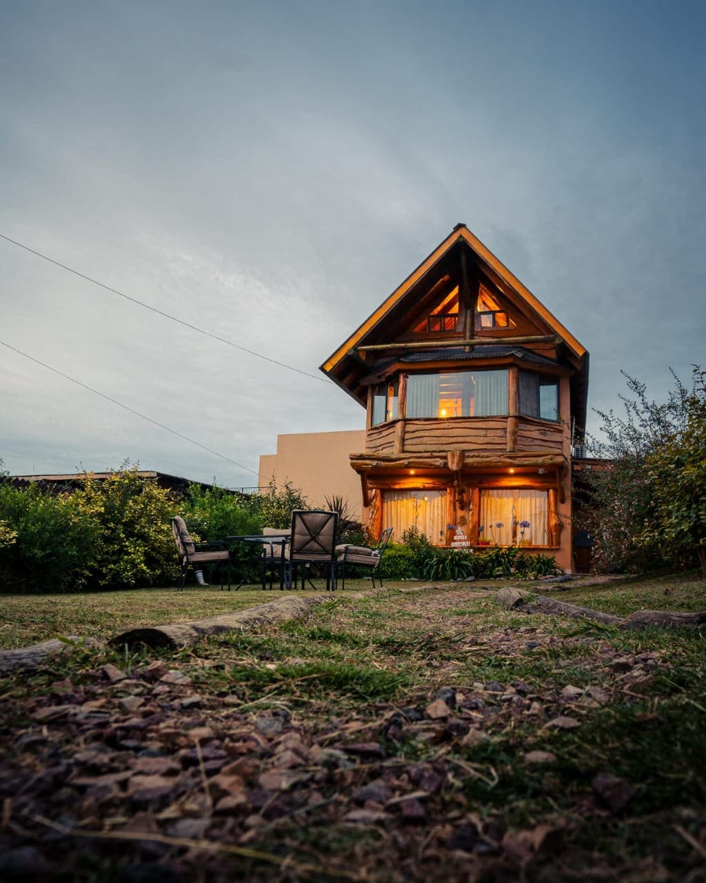 A charming cabin features an artistic wooden facade with large windows that illuminate the interior. A small outdoor seating area is visible, surrounded by landscaped greenery and rustic stone pathways, creating a welcoming outdoor space.