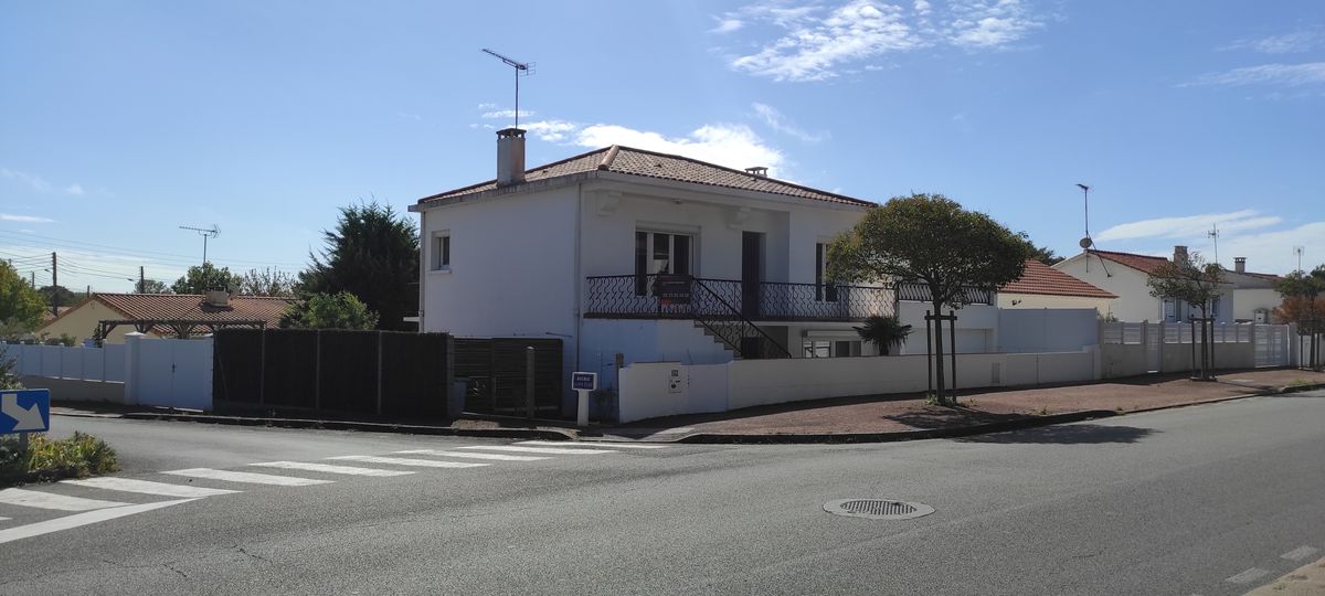 The exterior of the property is displayed, showcasing a two-story villa with a light-colored facade and a tiled roof. The building features a balcony, surrounded by a white fence and several trees. A street view includes nearby houses and open space.