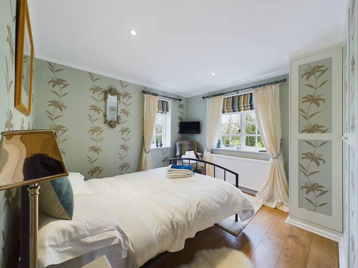 A calm bedroom features light blue wallpaper adorned with subtle floral patterns. A spacious bed with a white comforter is accompanied by a soft rug on wooden flooring. Natural light filters through decorated curtains, illuminating a small seating area and a mounted television.