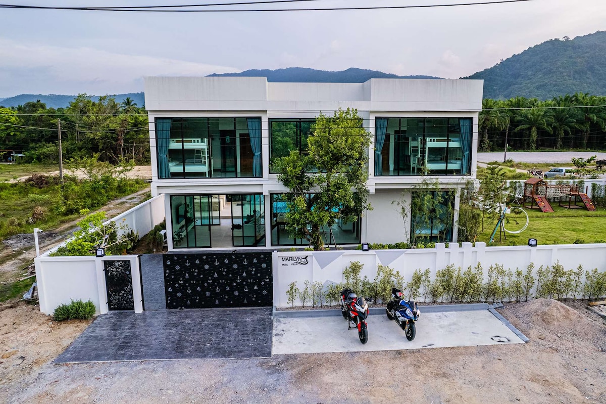 The exterior of a modern two-story house is depicted, featuring large glass windows and a clean architectural design. A spacious driveway accommodates two motorcycles. Lush greenery surrounds the property, complemented by the distant view of mountains and open landscapes.