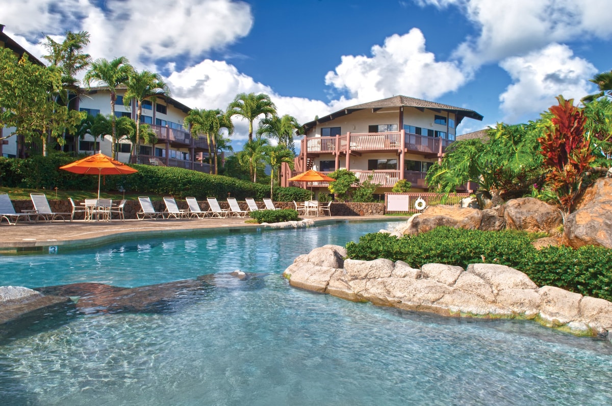 A clear swimming pool reflects the blue sky, surrounded by lush landscaping and tropical foliage. Lounge chairs with umbrellas are arranged by the pool, while the resort's buildings rise in the background, offering a harmonious blend of relaxation and nature.