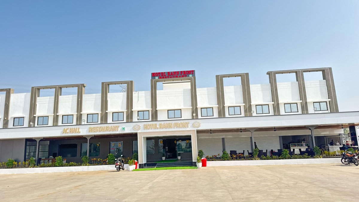 A modern hotel facade is presented, featuring a clear sky above. The entrance is marked by a sign, while the ground level includes a restaurant. Several outdoor seating options are visible, providing a welcoming space for guests.