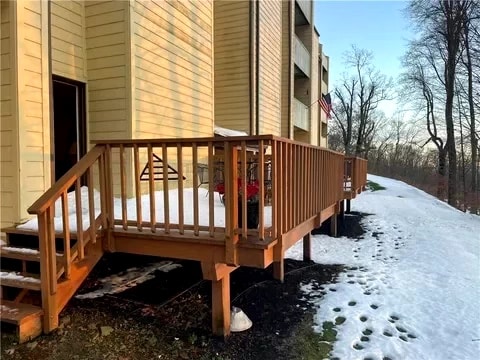 The exterior of the condo is depicted with a wooden ramp leading to the entrance, providing accessibility. A light dusting of snow covers the pathway and ground, while patches of bare earth are visible. Trees line the background, indicating a natural setting.