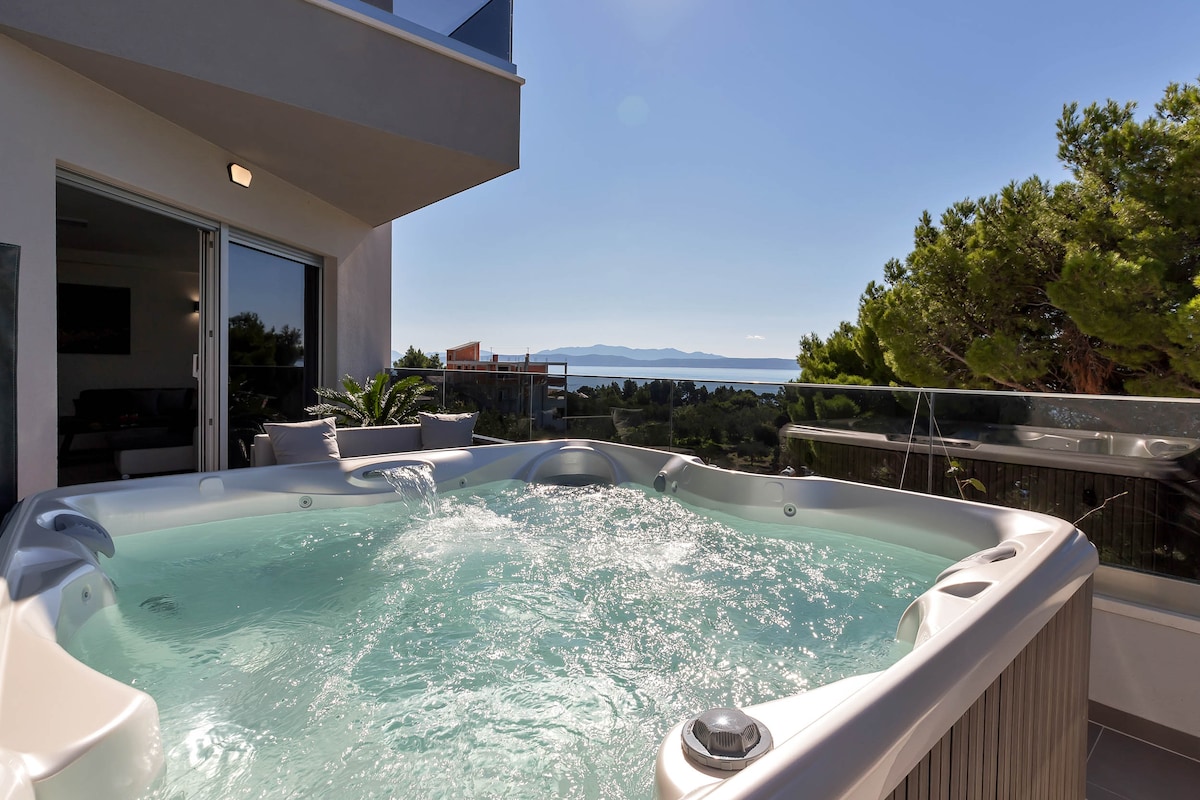 A private jacuzzi bubbles on the spacious terrace, framed by lush greenery. Stunning sea views are visible in the background, offering a serene setting for relaxation. Bright sunlight enhances the tranquil atmosphere, inviting guests to unwind in privacy.