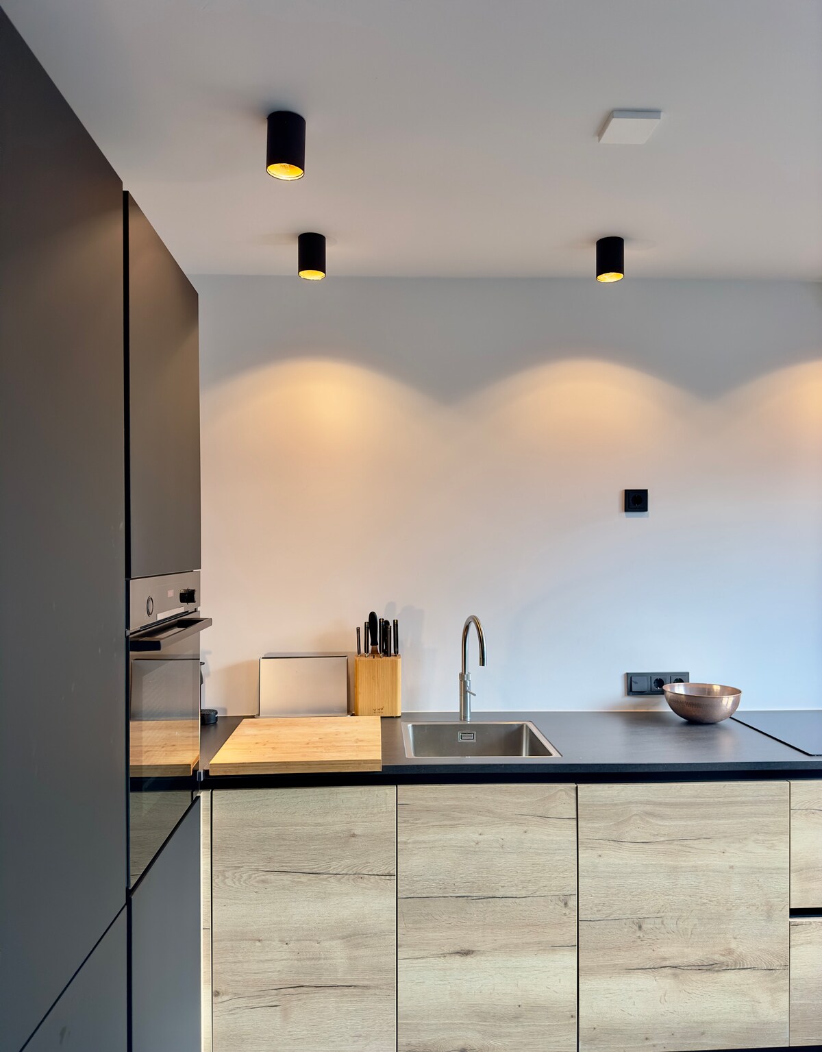 The kitchen area features sleek cabinetry in a combination of dark and light wood tones. A stainless steel sink is positioned under softly illuminated ceiling fixtures. Counter space is evident, complemented by kitchen utensils arranged neatly in a wooden holder.